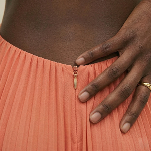 Person wearing a coral pleated skirt with a gold zipper and hand adjusting it.