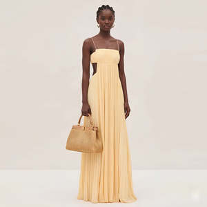 Woman in a long beige dress holding a woven handbag against a plain background