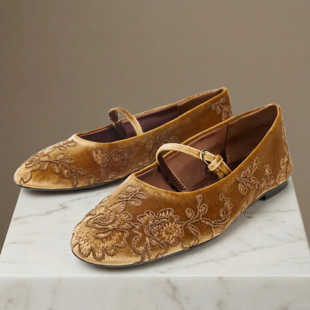 Pair of brown velvet shoes with floral embroidery on a marble surface.