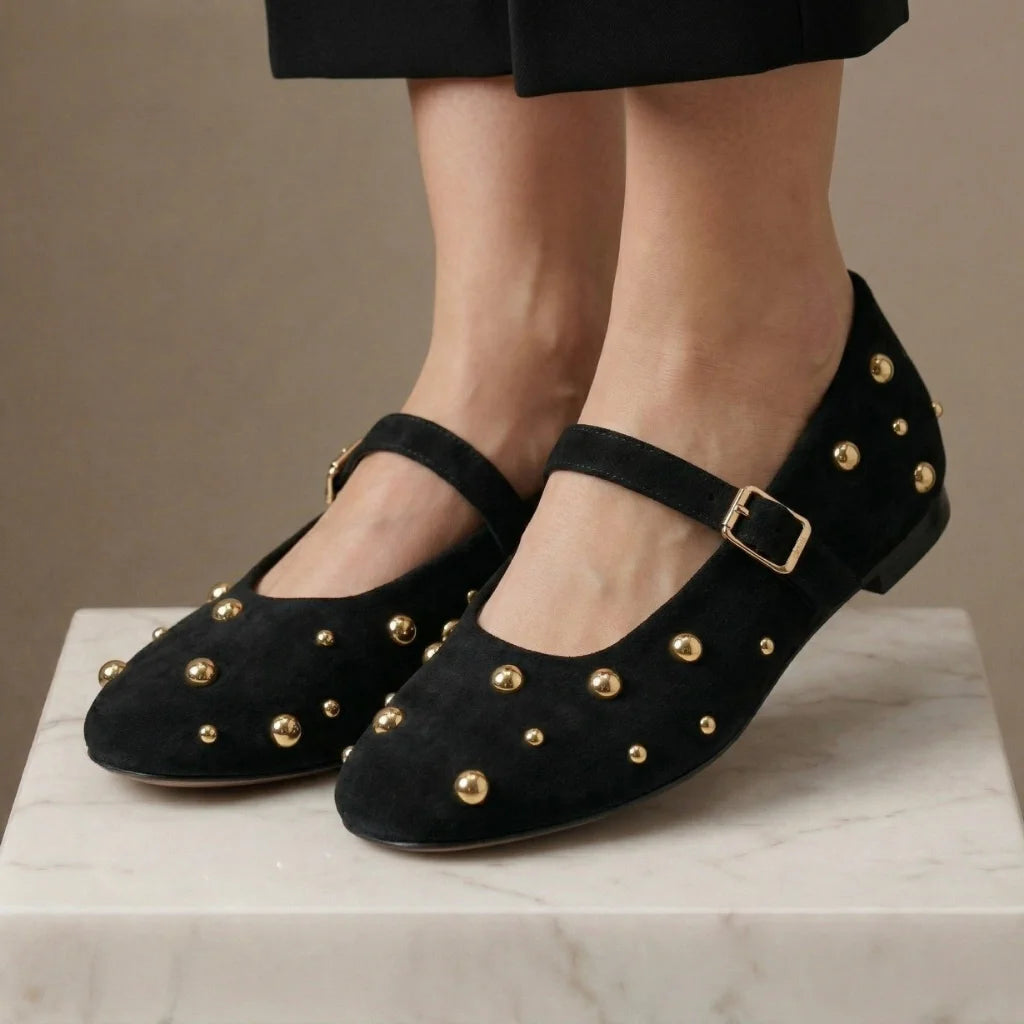 Black suede shoes with gold studs and buckles worn by a person on a marble surface.
