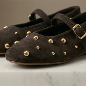 Brown suede shoes with gold studs and buckles on a marble surface.