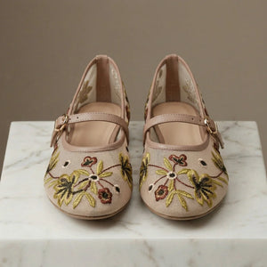 Pair of beige shoes with floral embroidery on a marble surface.