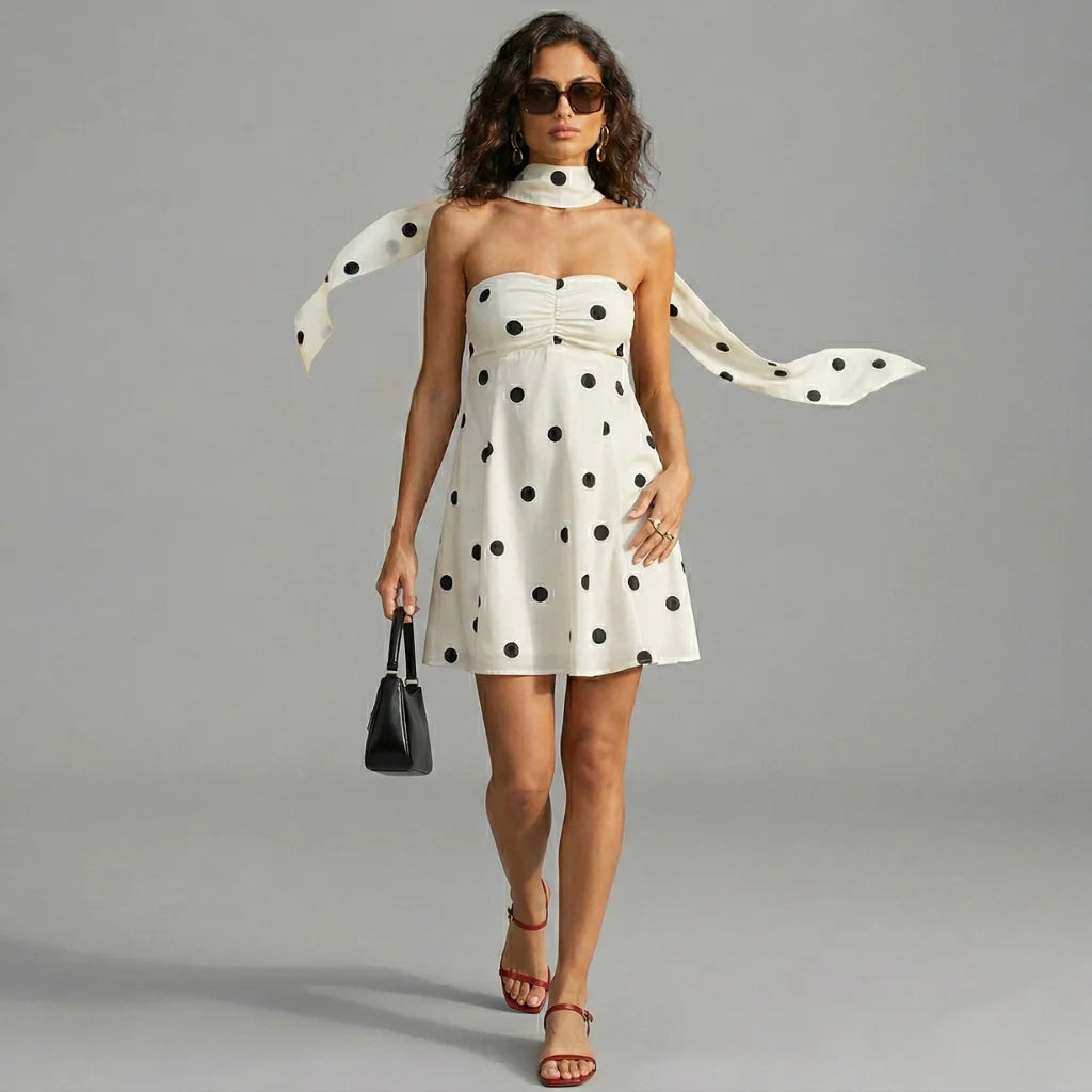 Woman wearing a polka dot dress with a scarf, holding a handbag, on a gray background