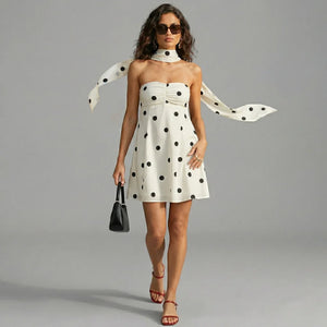 Woman wearing a polka dot dress with a scarf, holding a handbag, on a gray background