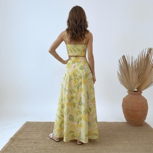 Woman wearing a yellow floral dress standing on a woven mat with a vase of dried plants in the background.