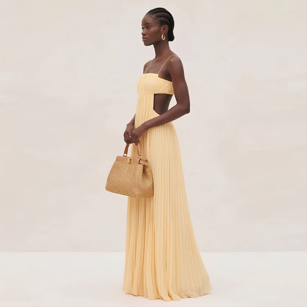 Woman in a long yellow dress holding a woven handbag against a plain background