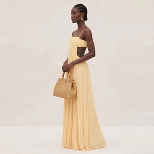 Woman in a long yellow dress holding a woven handbag against a plain background