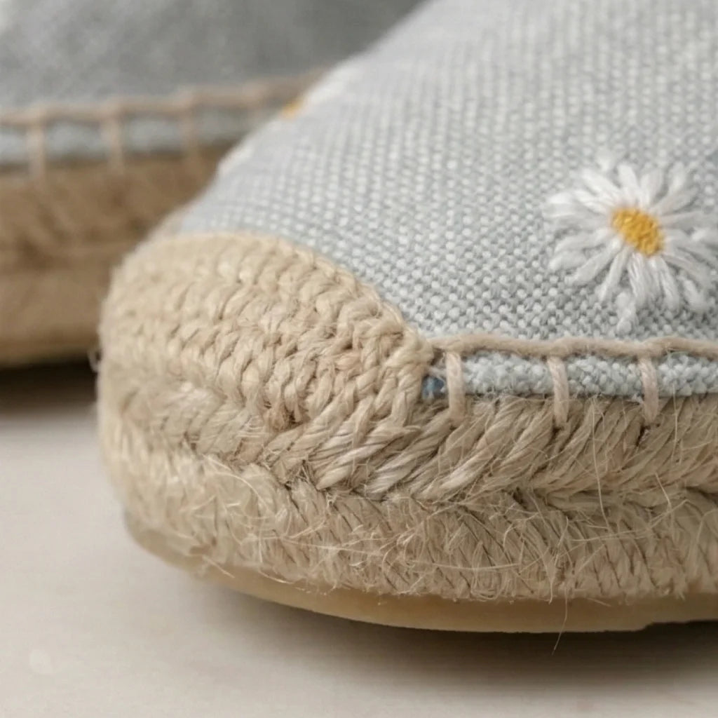 Close-up of a light gray espadrille shoe with jute sole and embroidered daisy on a neutral background.