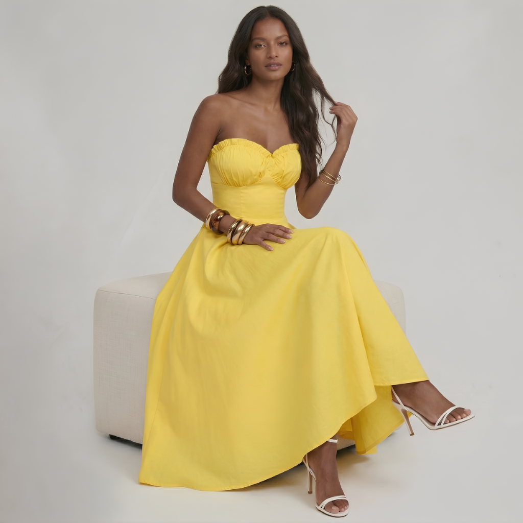 Woman in a yellow strapless gown sitting on a white chair against a plain background